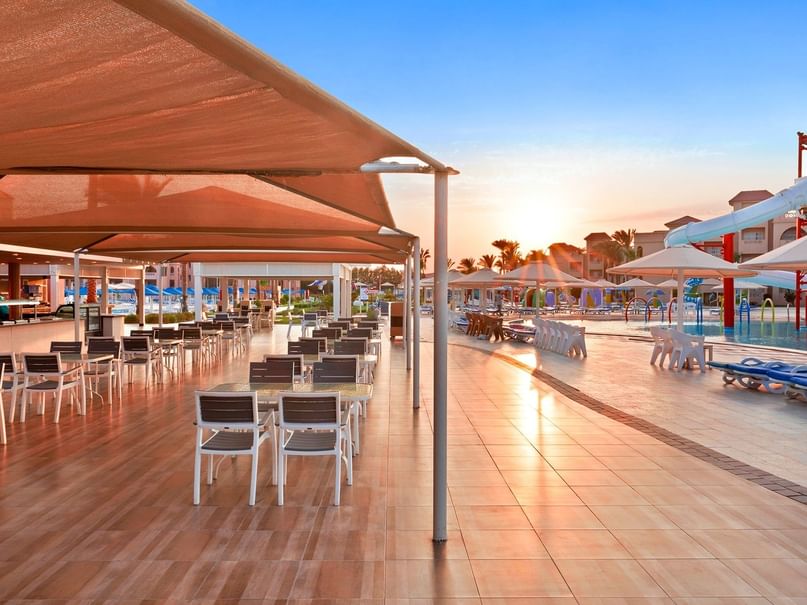 Outdoor dining area with tables and chairs under canopy, poolside with umbrellas and water slides at sunset.