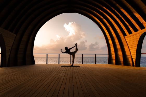 Silhouette of a woman in a yoga pose under a wooden archway at sunset, overlooking the ocean at Ananea Madivaru Maldives
