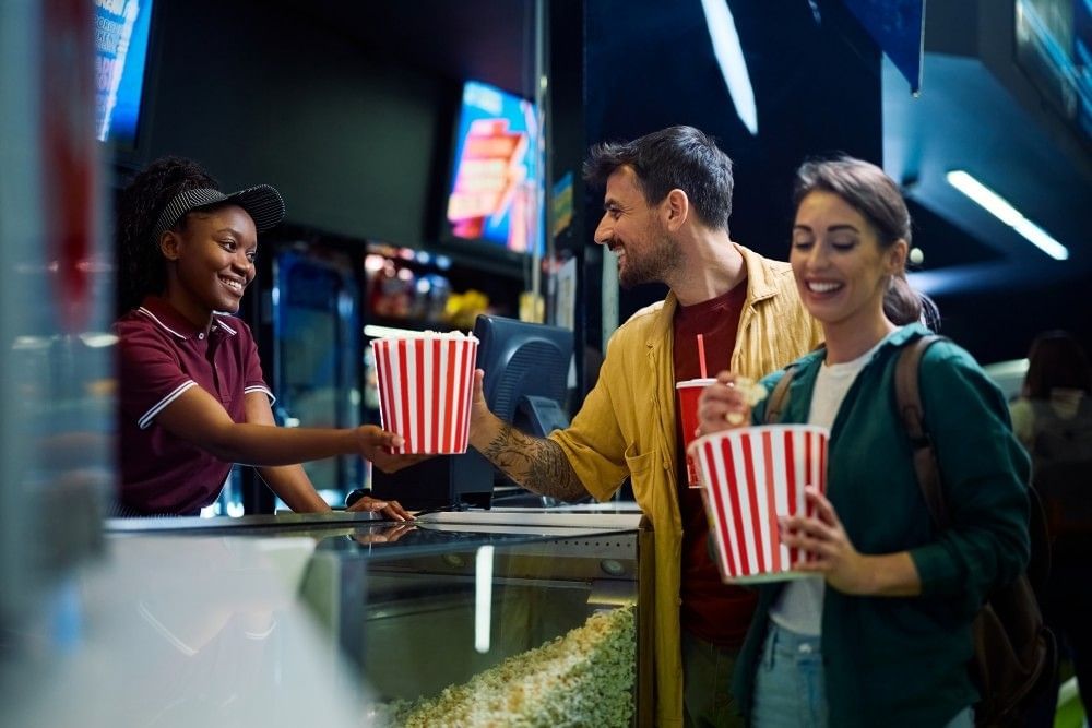 A man and woman take popcorn buckets and sodas from a cashier at a movie theatre concession counter. 