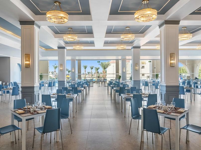 An empty restaurant with blue chairs and white tables, large windows, and decorative ceiling lights.
