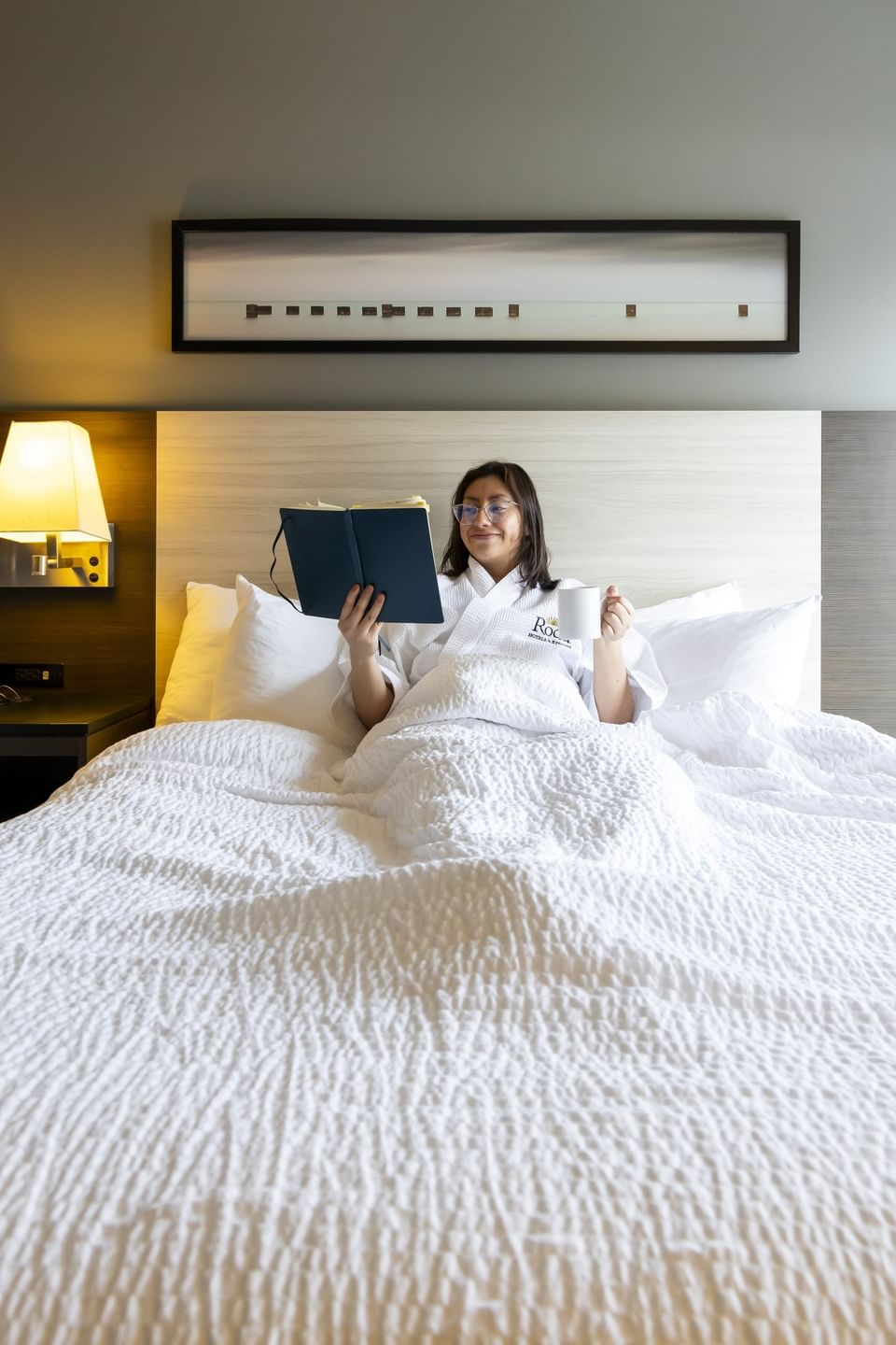 Guest reading a book in bed under a soft wall lamp, with a scenic frame surrounding it at Rodd Miramichi River