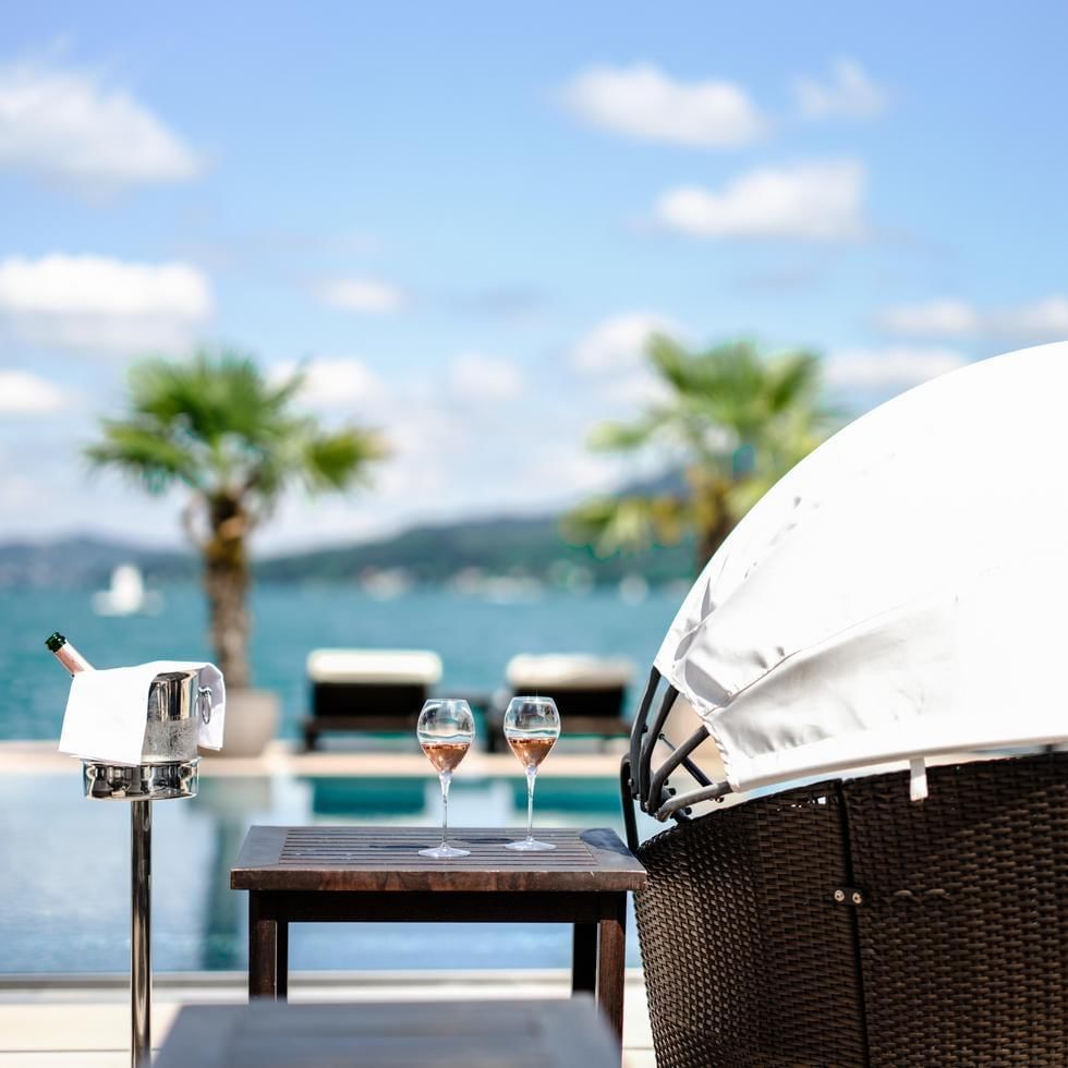 Poolside lounge with wine glasses, bottle, and canopy on deck overlooking water and palm trees.