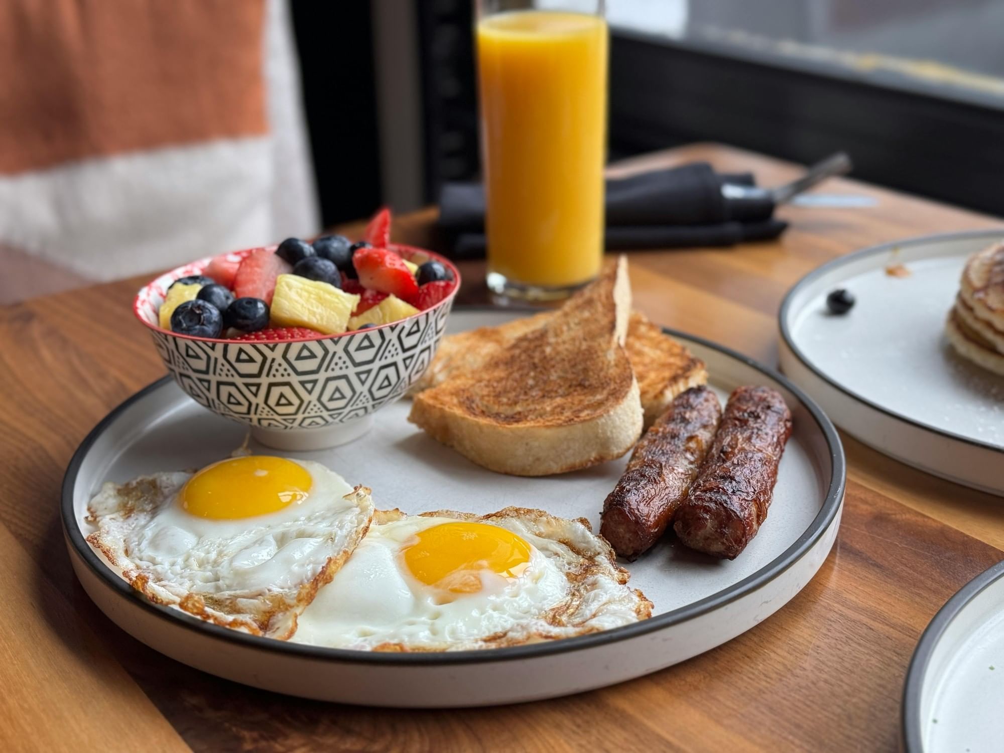 Breakfast plate with fried eggs, sausages, toast, and a fruit bowl served with orange juice at Amora Hotel Adelaide