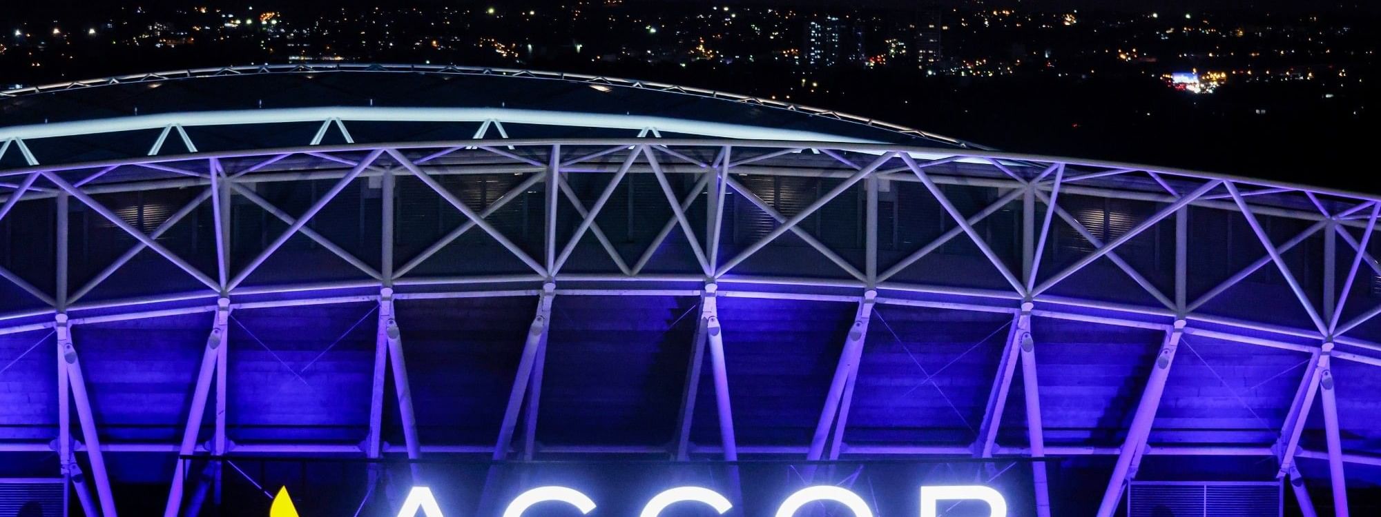 Accor Stadium illuminated with purple lighting and bright signage against an orange sunset sky with city lights in background.