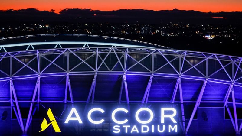 Accor Stadium illuminated with purple lighting and bright signage against an orange sunset sky with city lights in background.