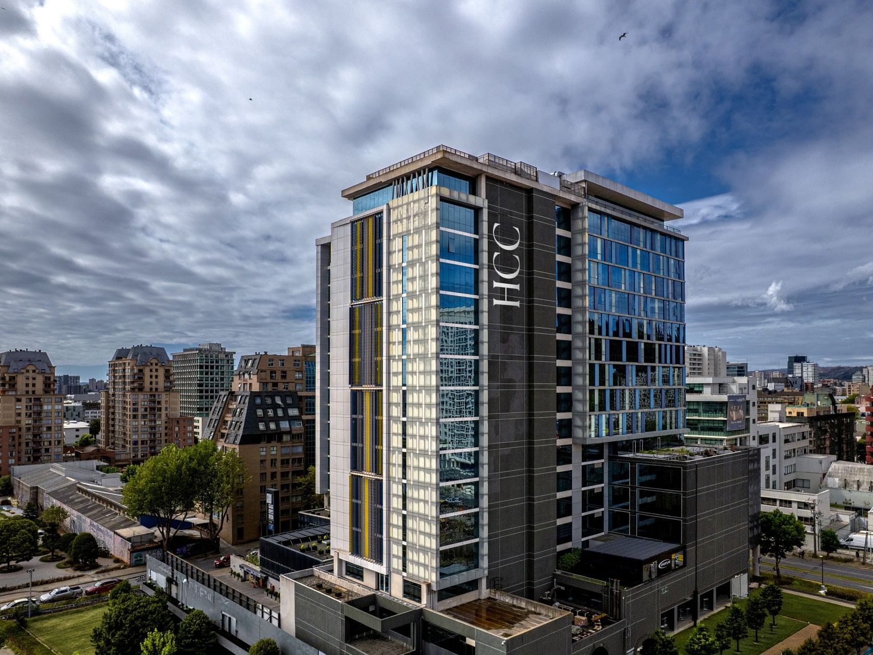 Un edificio alto y moderno de un hotel, con fachada acristalada y el logotipo de HCC, entre otros edificios y zonas verdes.