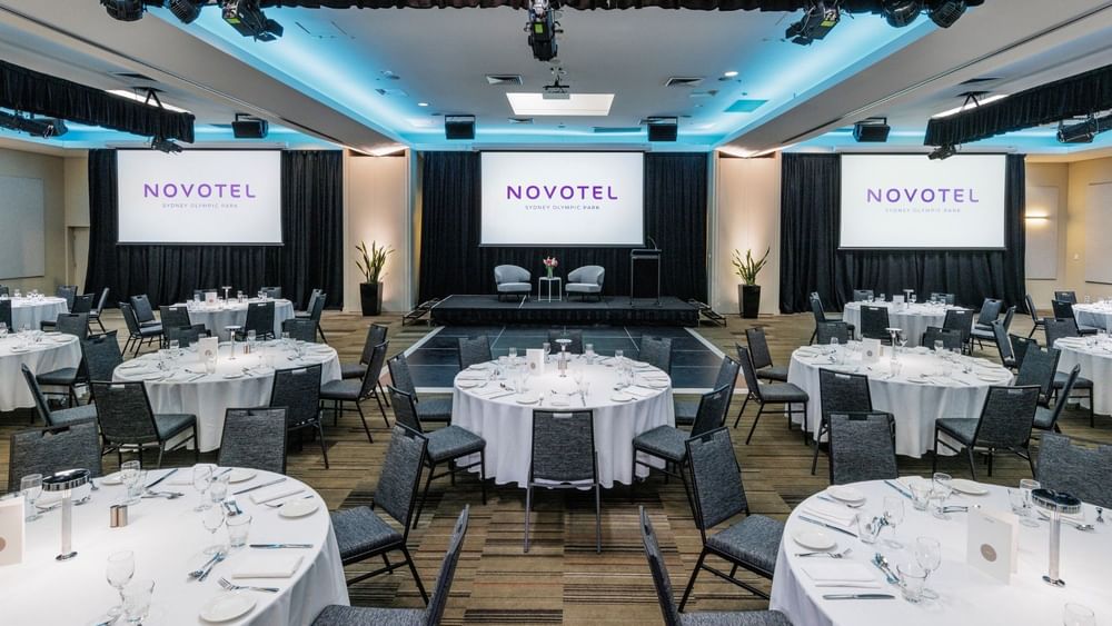 Modern event space with round tables, stage, blue lighting, and branded screens