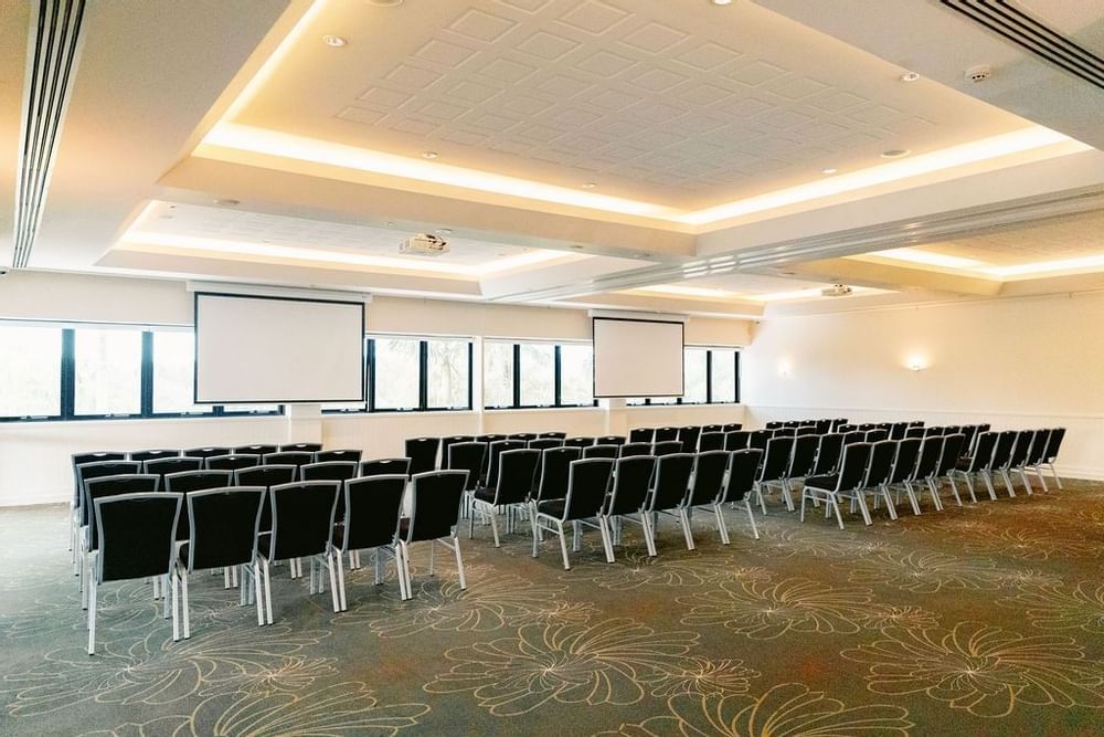 Empty conference room with rows of chairs, two projectors, and large windows with black frames.