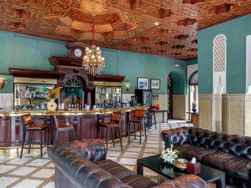 Elegant bar with ornate ceiling, leather couches, and a central bar counter with stools and a chandelier.