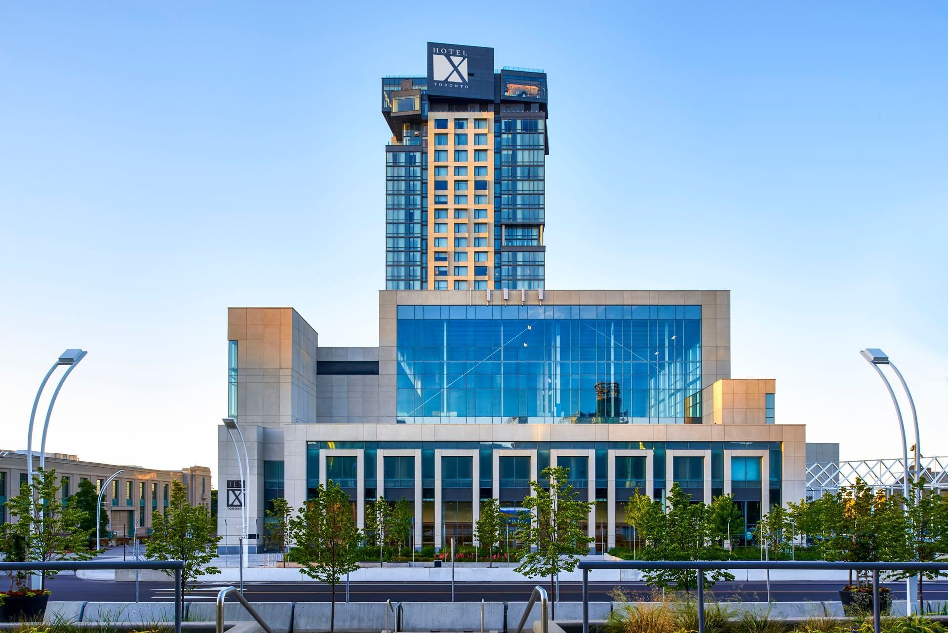 Modern glass tower hotel exterior facing street lined with young trees and clear blue sky at Hotel X Toronto Library