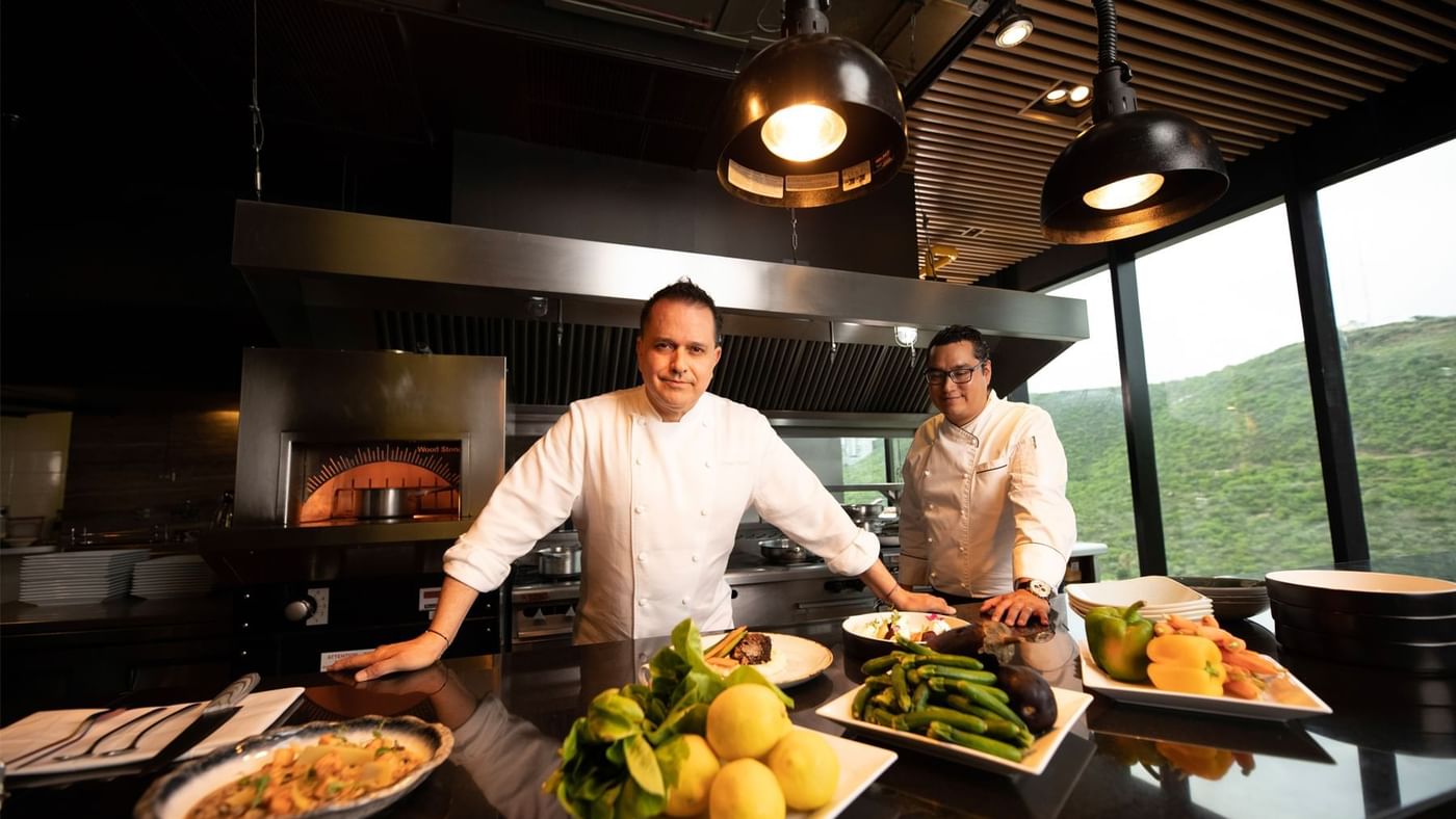 Two chefs stand in a modern kitchen beside an array of fresh ingredients and beautifully plated dishes at Live Aqua