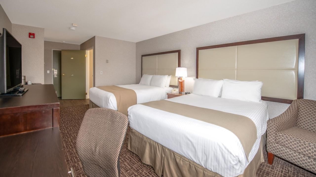 Hotel room with two beds, nightstand with lamp, TV, and two chairs.