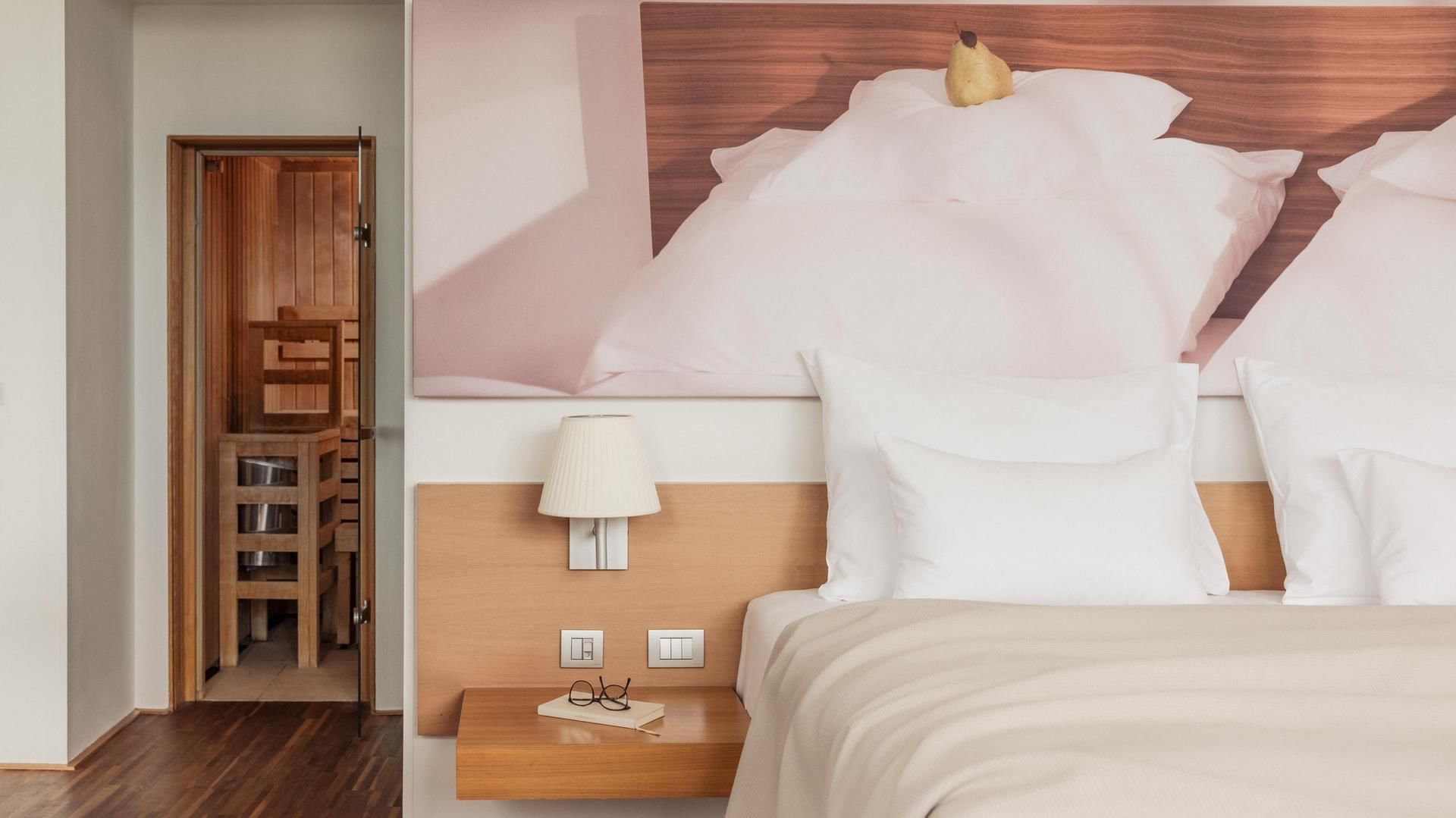 Hotel room with bed, wall art, lamp, wooden nightstand, eyeglasses, and open door to another room.