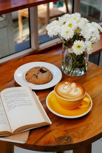 A small wooden table set with an open book, a vase of white flowers, a cookie, and a cappuccino. 