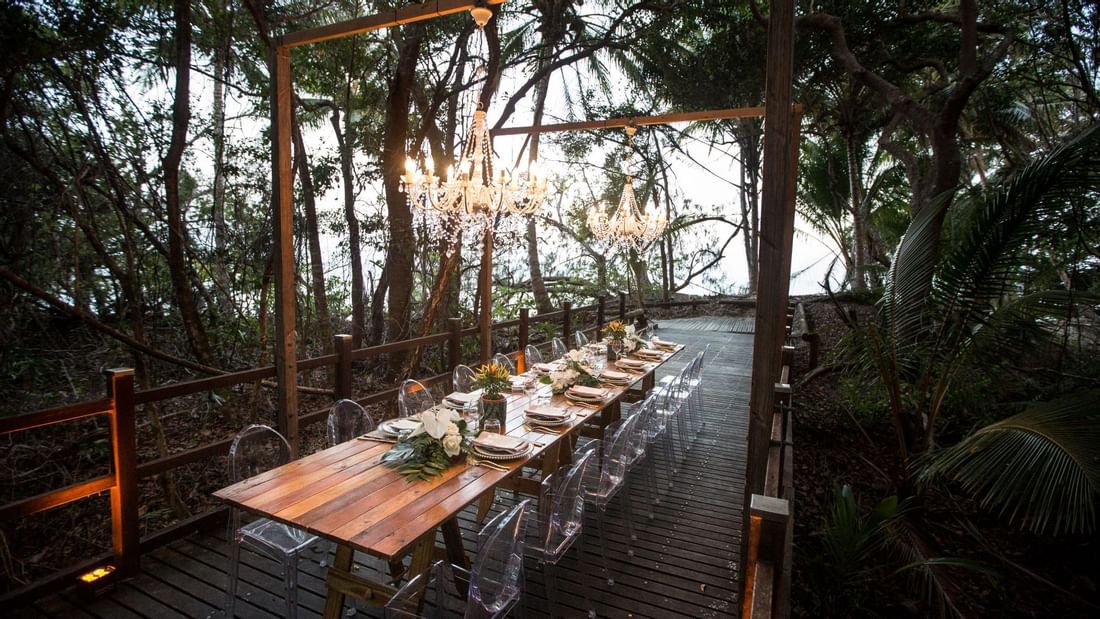 Elegant outdoor dining setup with chandeliers, long wooden table & floral centerpieces in Boardwalk at Pullman Port Douglas