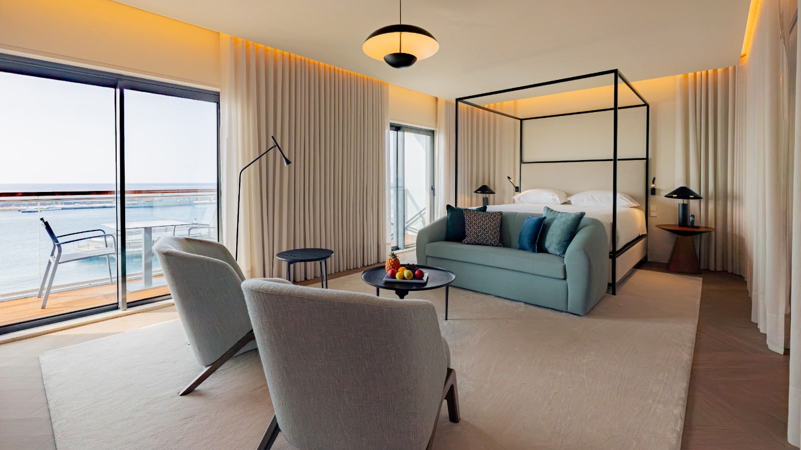 Quarto de hotel com cama, sofá, mesa, cortinas e vista para o mar.