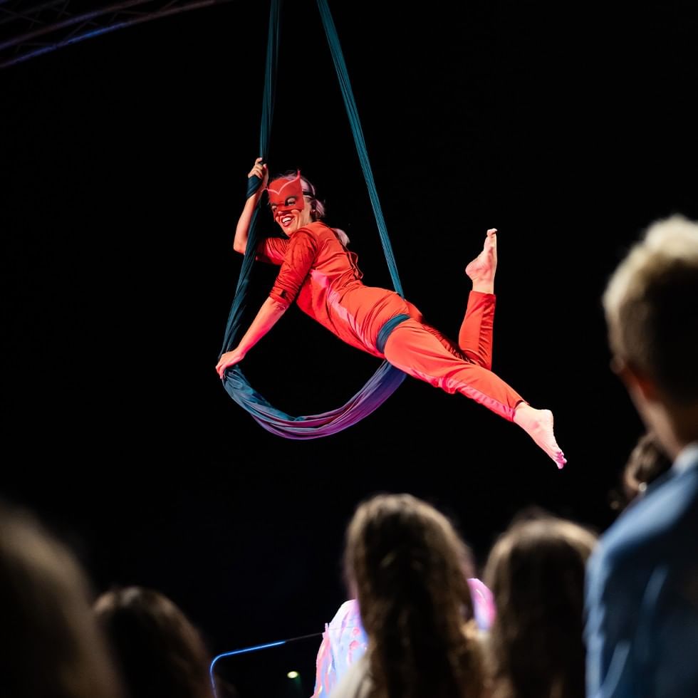 Acrobata in costume rosso e maschera rossa sospesa su un'amaca blu davanti a un pubblico.