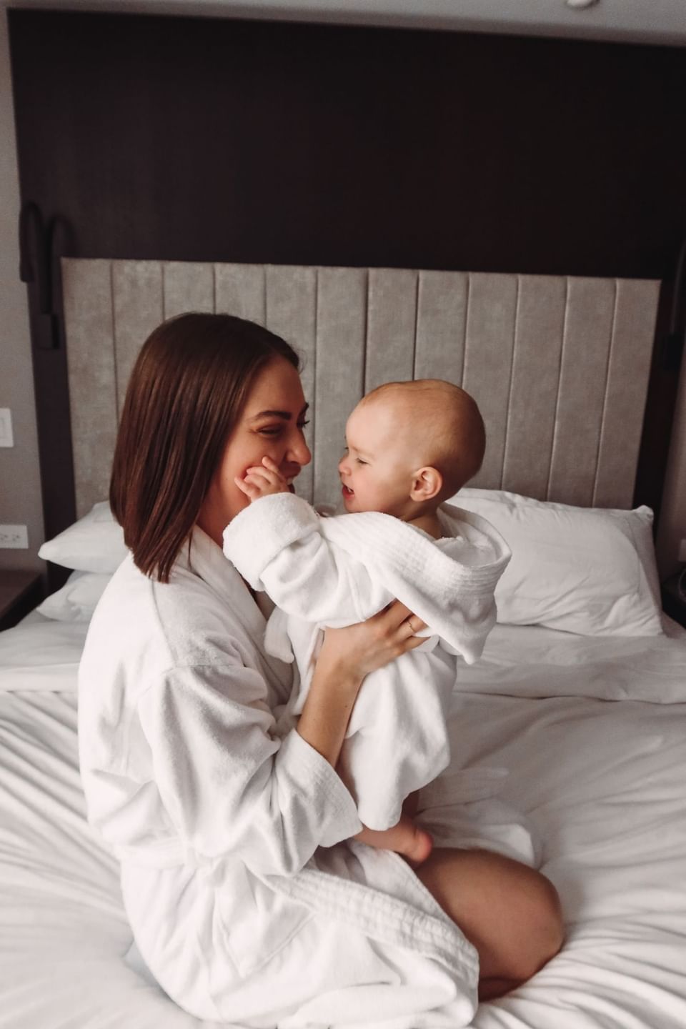 Woman in white bathrobe holding a baby on plush white bed with upholstered headboard in bedroom suite at Hotel X Toronto