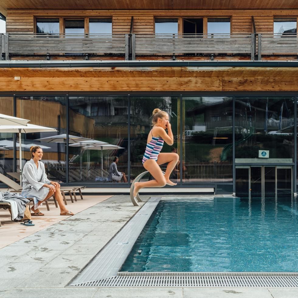 Girl jumping into pool at resort, woman in robe watching, modern building with large windows.