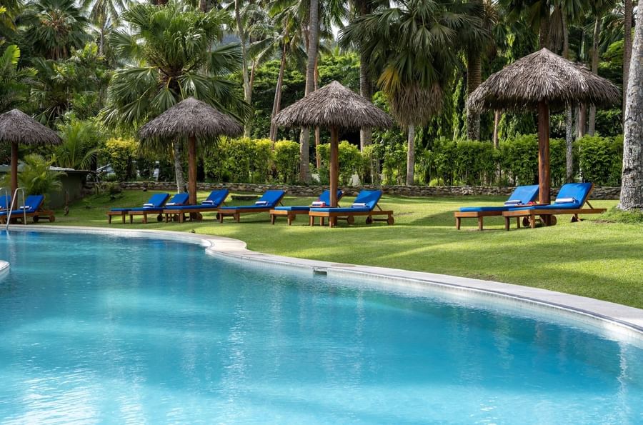 Blue lounge chairs by thatched umbrellas on a green lawn under palms near the pool at Tambua Sands Beach Resort
