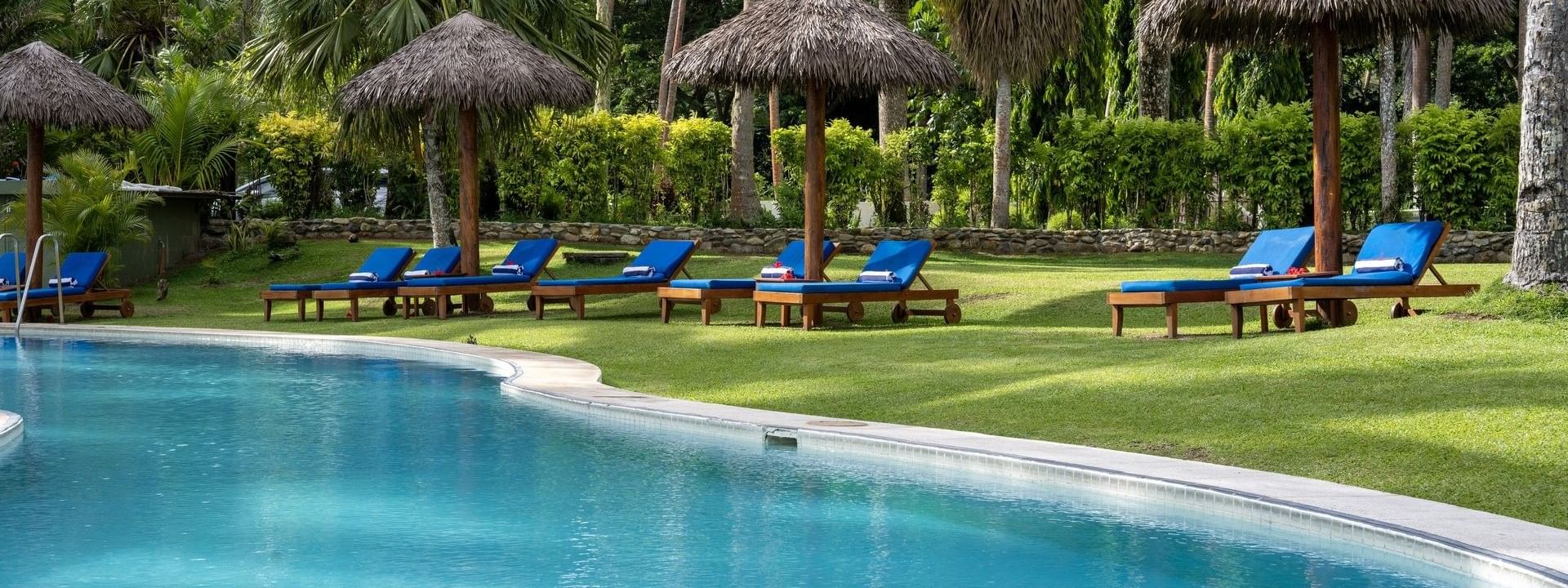 Blue lounge chairs by thatched umbrellas on a green lawn under palms near the pool at Tambua Sands Beach Resort