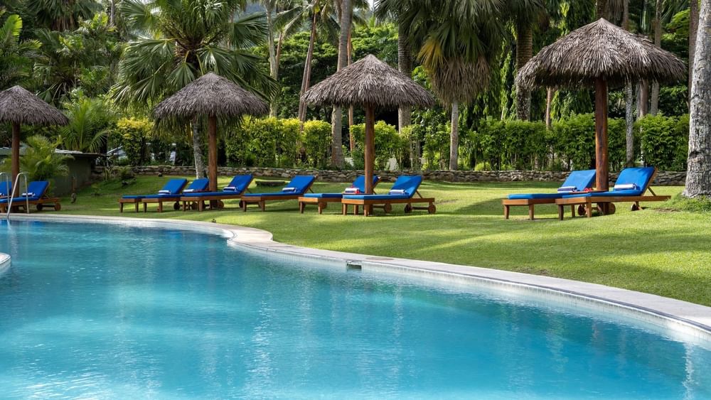 Blue lounge chairs by thatched umbrellas on a green lawn under palms near the pool at Tambua Sands Beach Resort