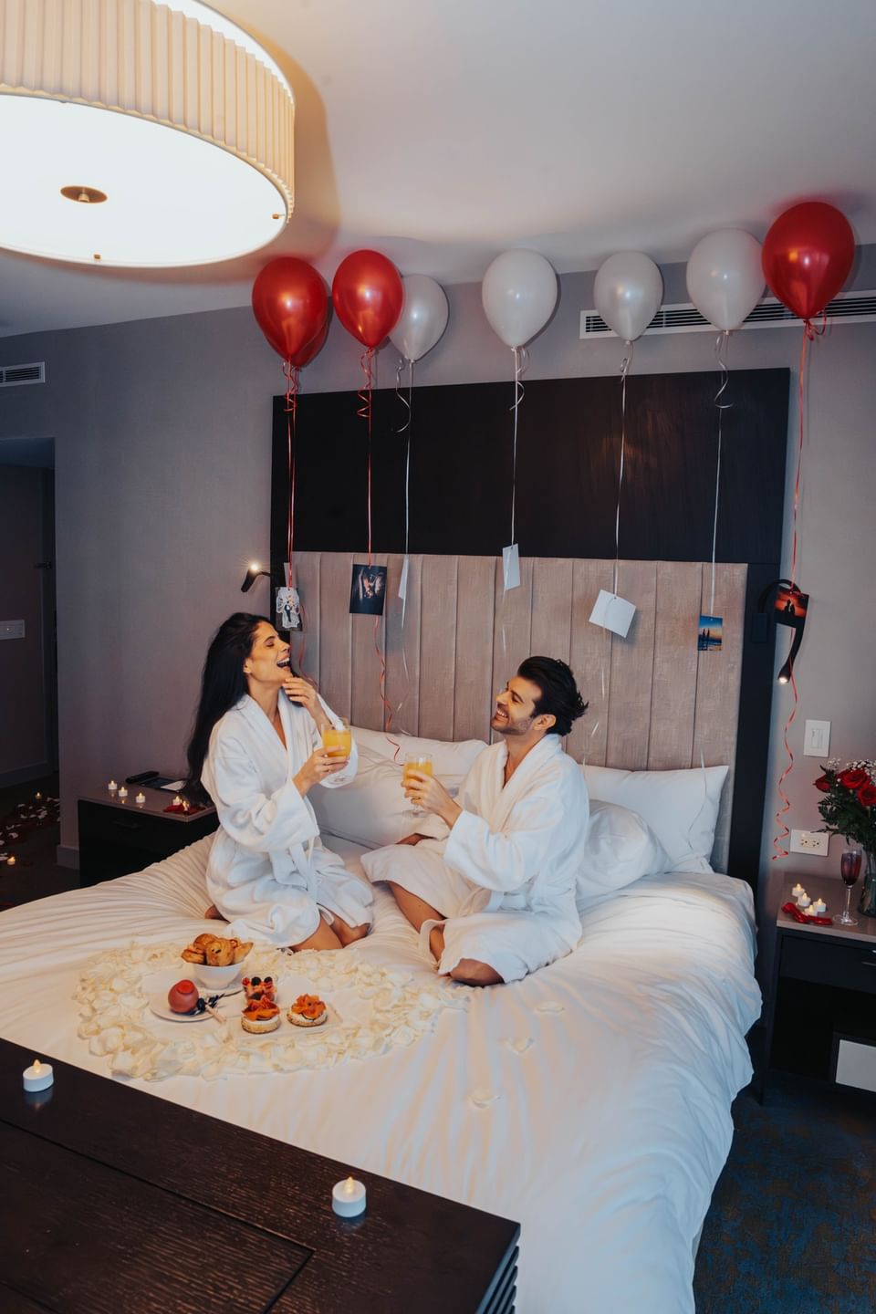 Couple sharing mimosas on a bed, under red and white balloons with photos hanging above at Hotel X Toronto