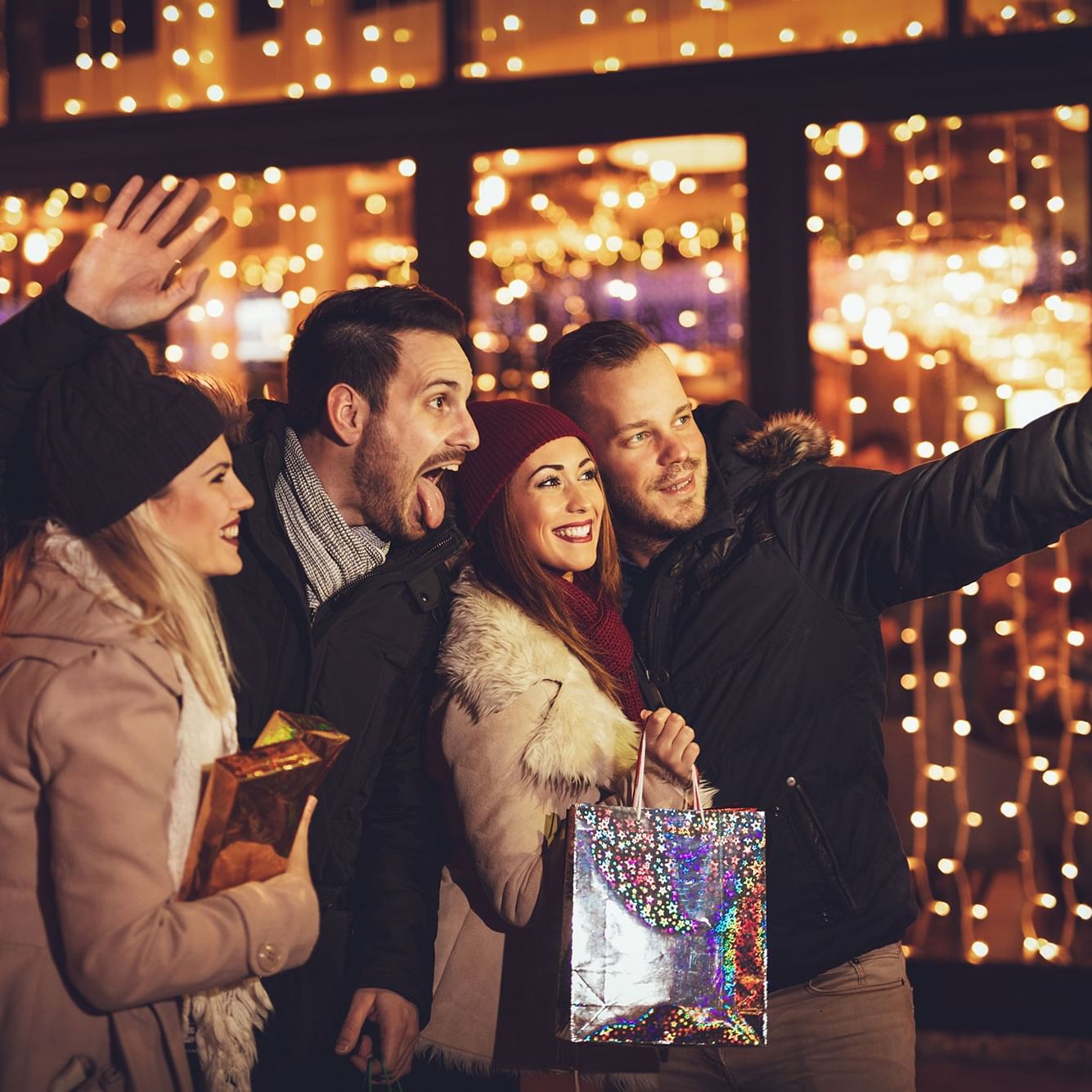 Four friends with shopping bags taking a selfie against a festive string light backdrop.