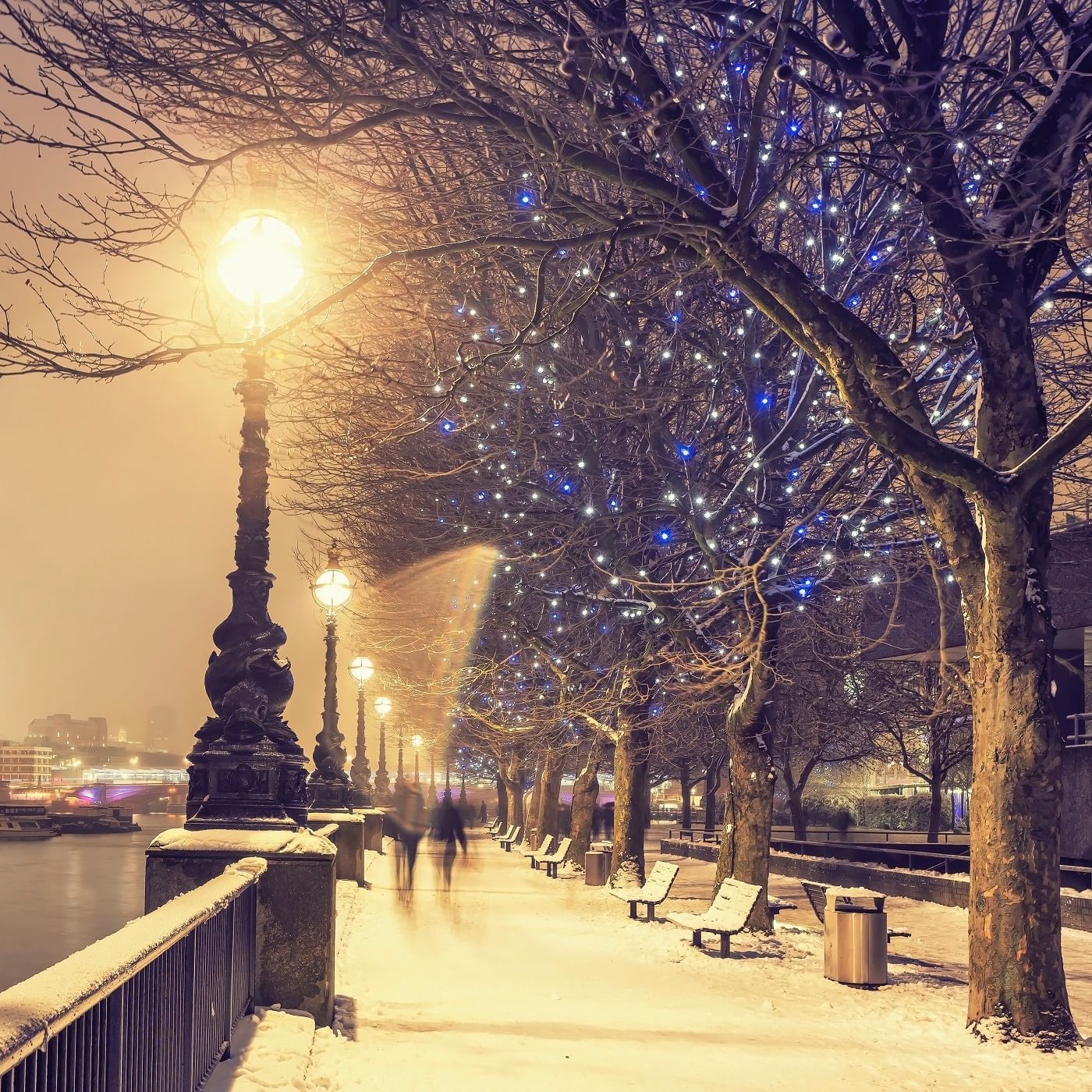 Snow-covered riverside promenade adorned with sparkling blue and white lights, creating a festive scene near The Londoner