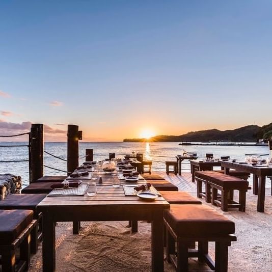 A beachside dining setup with wooden tables and benches, set against a sunset backdrop with a clear view of the ocean.