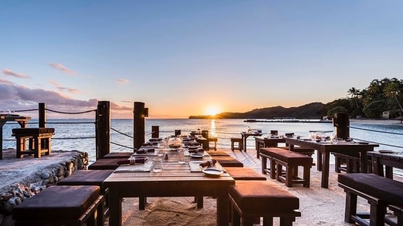 A beachside dining setup with wooden tables and benches, set against a sunset backdrop with a clear view of the ocean.
