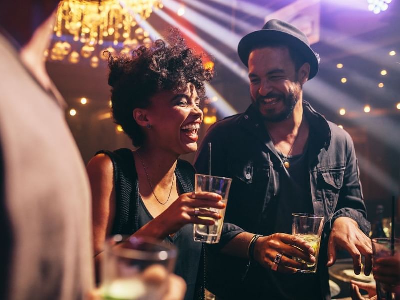 Dos amigos riendo y disfrutando bebidas bajo luces coloridas en el animado club nocturno Fiesta Americana Funeeq