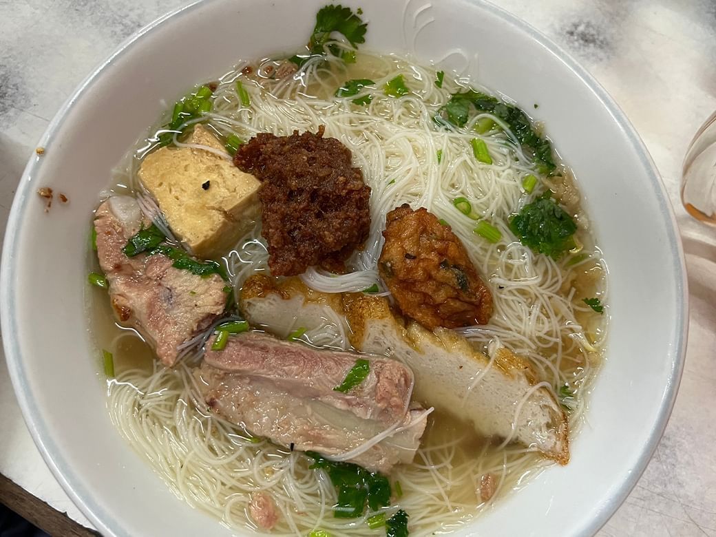 A white bowl of noodle soup with duck tofu and herbs at Hop Inn Hotel on a table