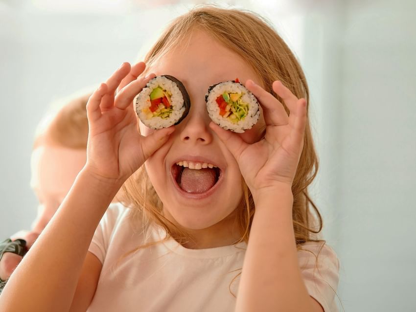 A young girl smiling and holding two sushi rolls over her eyes like goggles at Camino Real Hotel