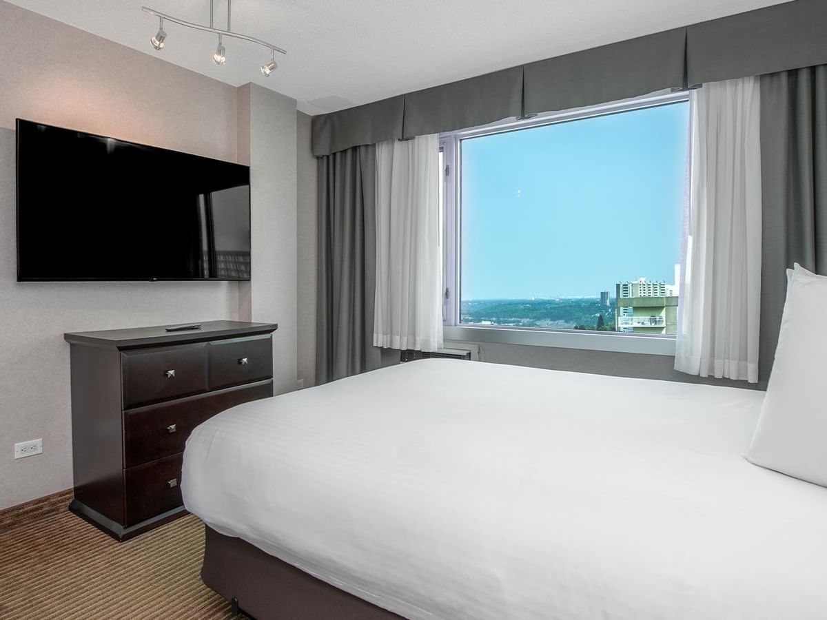 Hotel guest suite with a queen bed, wall-mounted TV, dresser and large window with a city view.