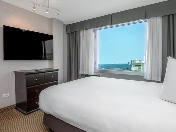 Hotel guest suite with a queen bed, wall-mounted TV, dresser and large window with a city view.