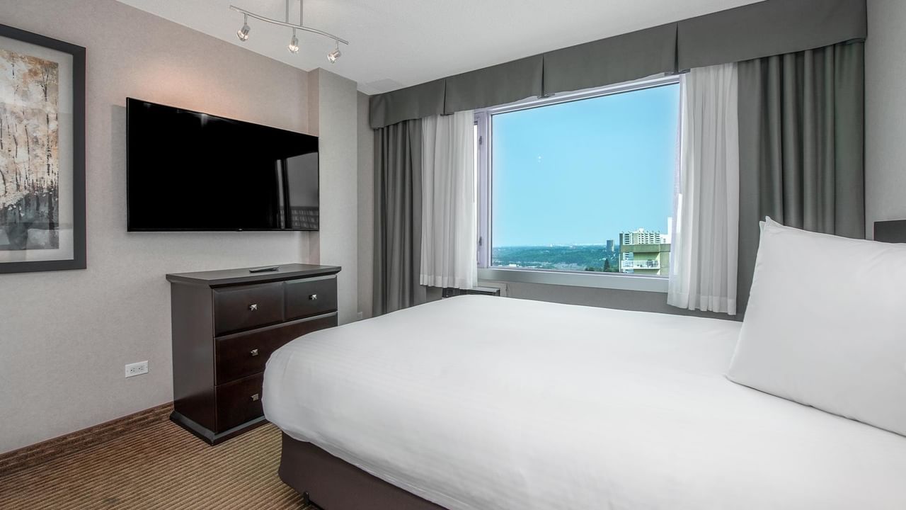 Hotel guest suite with a queen bed, wall-mounted TV, dresser and large window with a city view.