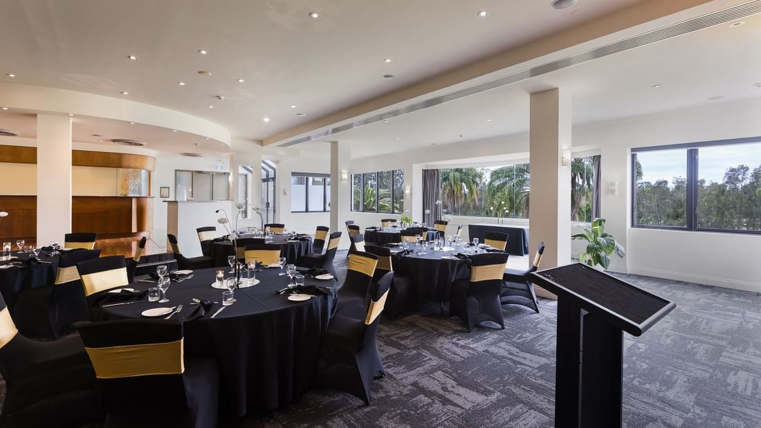 A banquet hall with round tables, black chairs with yellow sashes, a podium, and large windows showing trees outside.