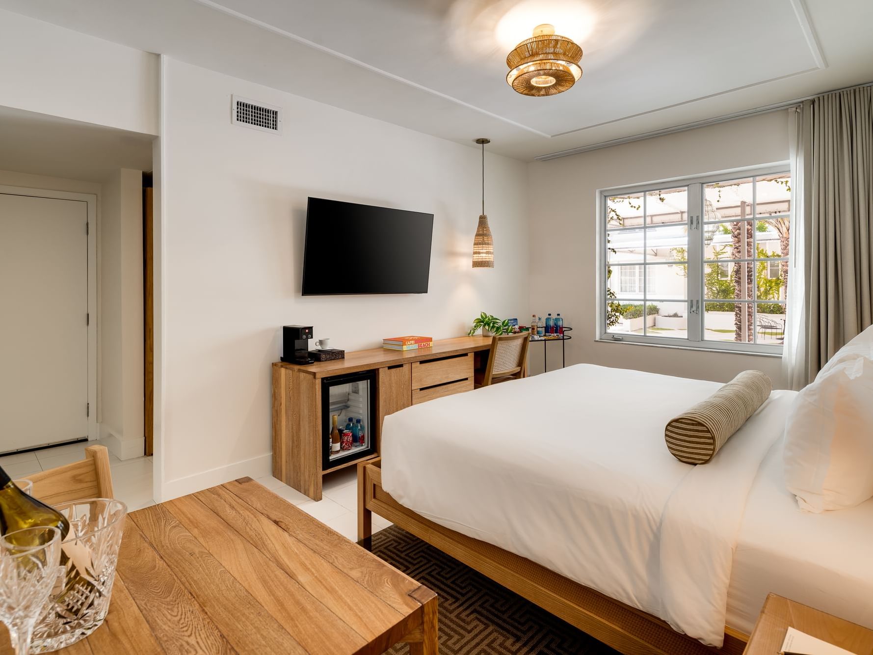 Hotel room with bed, TV, refrigerator, desk, wine, and window with curtains and outdoor view.