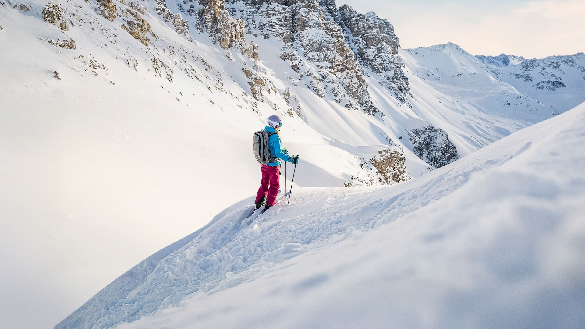 A skier with backpack and poles navigates a snowy mountain slope with rugged terrain.