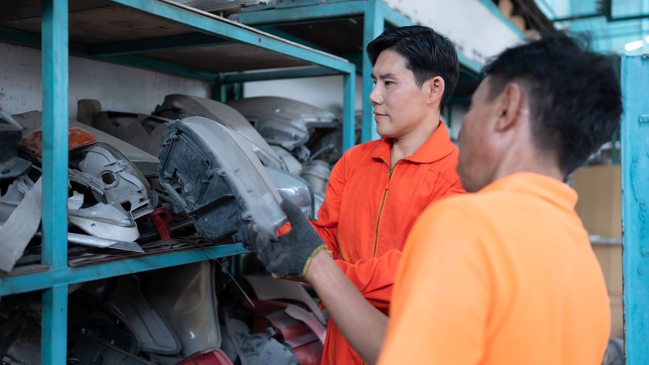 Two men in orange shirts examining automotive parts on blue shelving at an Early Bird Swap Meet
