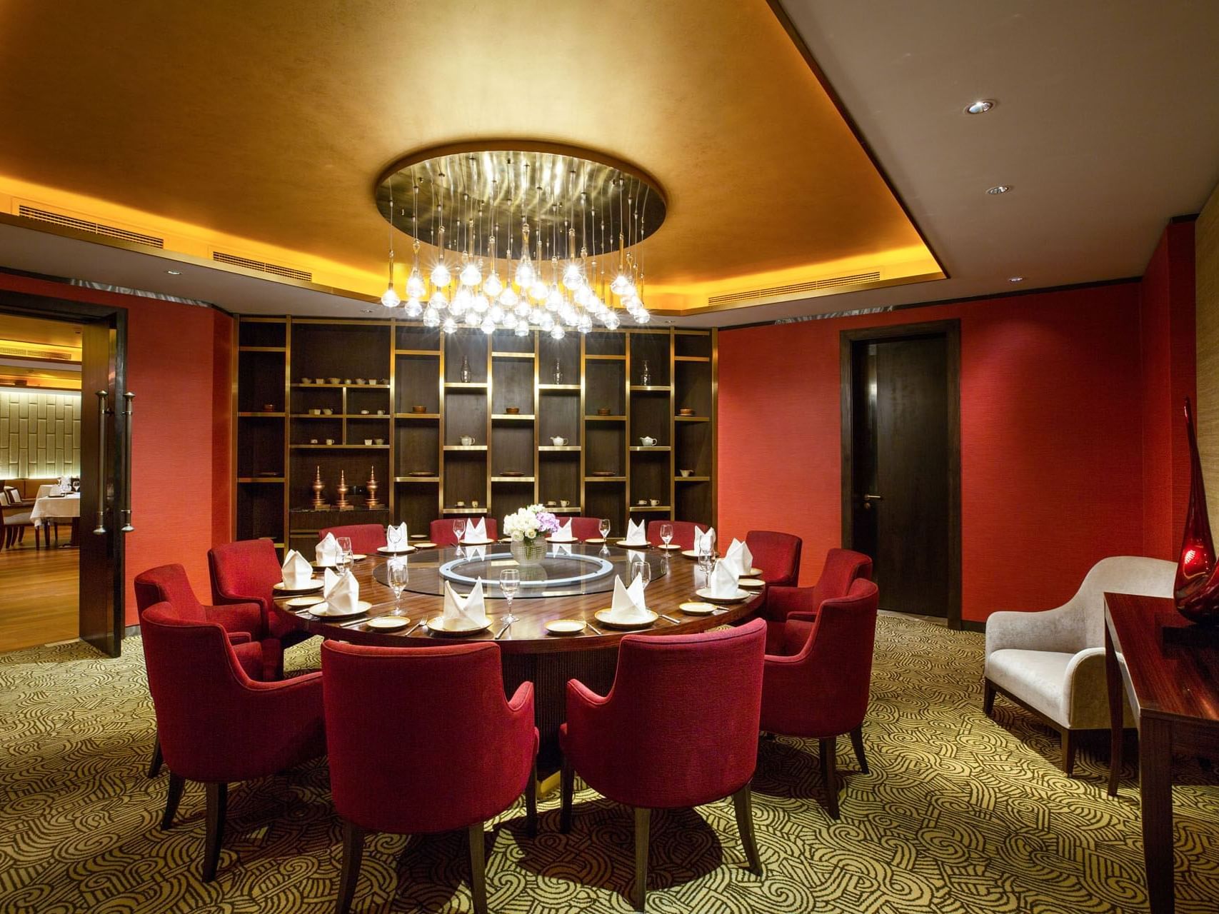 Elegant dining room with round table, red chairs, chandelier, red walls, and patterned carpet.