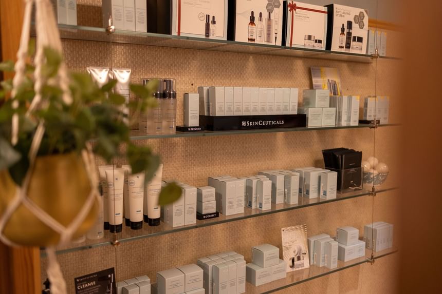 A glass display shelf with various skincare products and a hanging plant, with a blurred background.