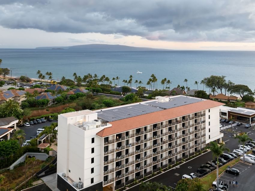A large white building with a red roof and balconies, surrounded by a parking lot and palm trees, with a view of the ocean in the background.