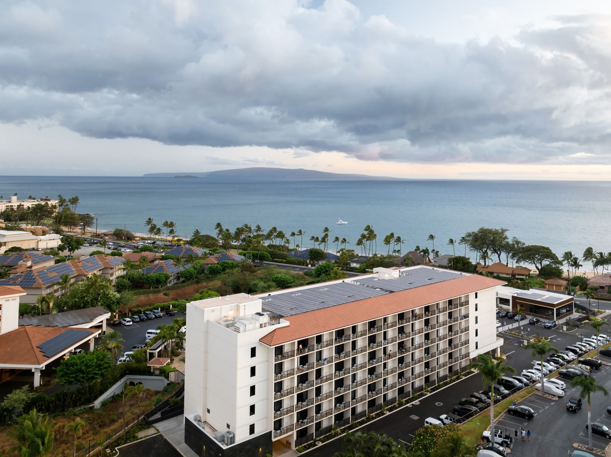 A large white building with a red roof and balconies, surrounded by a parking lot and palm trees, with a view of the ocean in the background.
