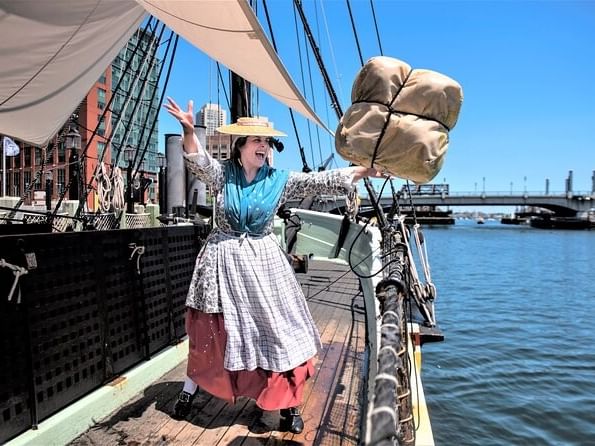 Woman in period clothing on a ship deck holding a large bag, with water and bridge in the background.