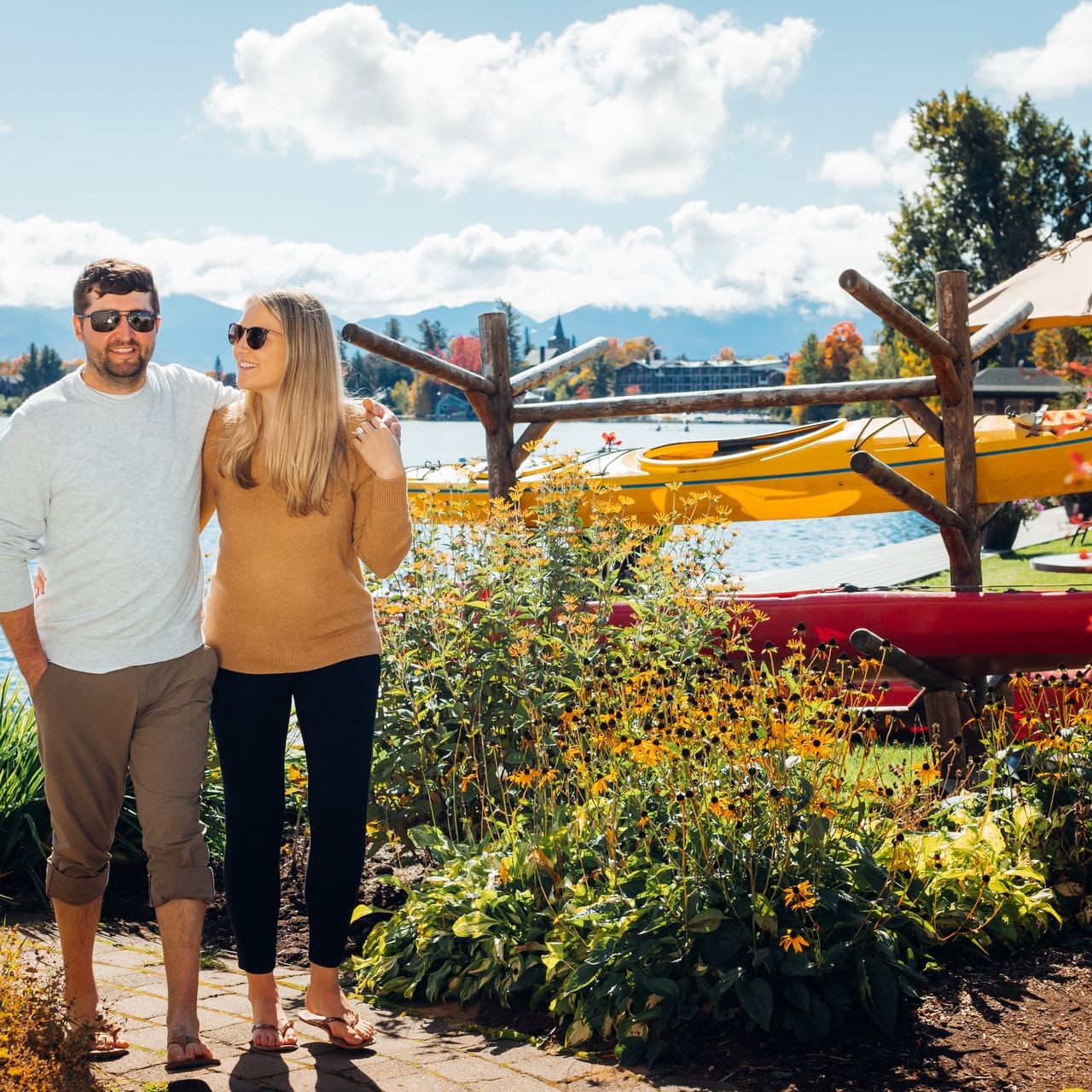Couple stands by lake with yellow and red kayaks, trees, and mountains in background.