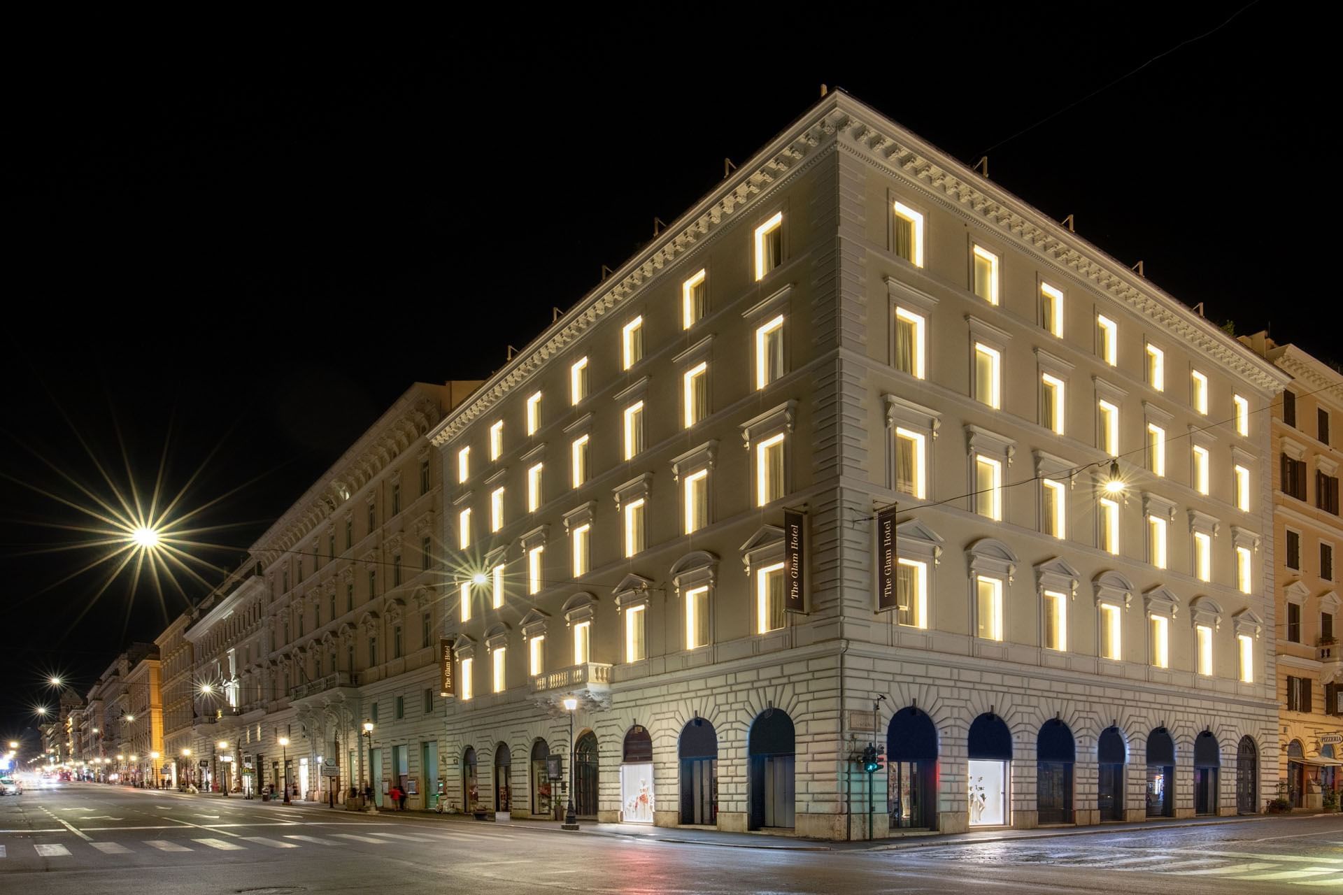 The Glam Hotel facade illuminated at night, showcasing bright windows and classical architecture on a quiet street