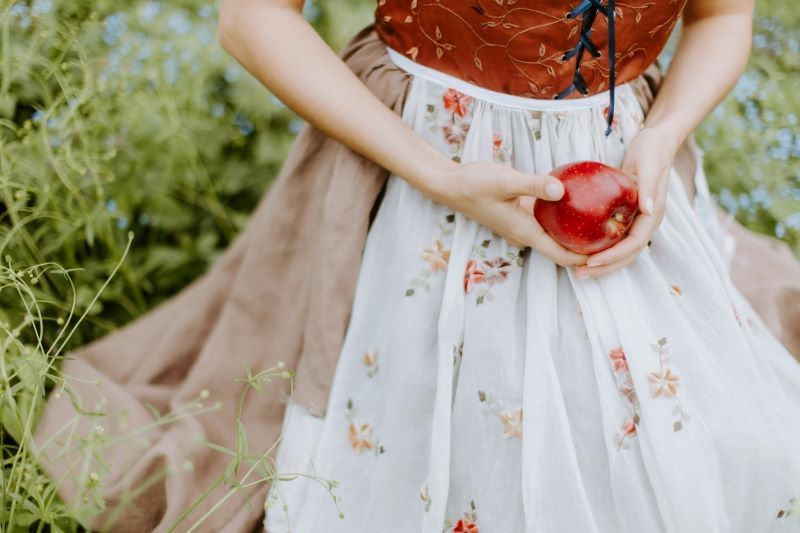 An image of a woman neck down sitting in grass, wearing a red top, beige skirt, and white hip apron, while holding an apple.