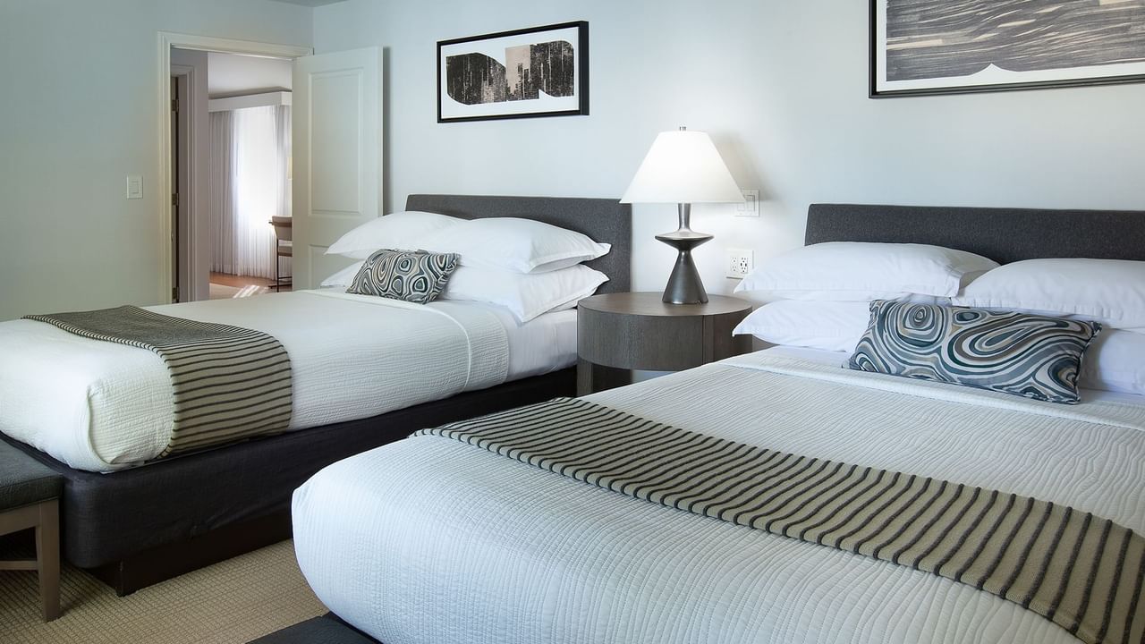 Two beds with pillows and throws, nightstand with lamp, and framed art on white walls.